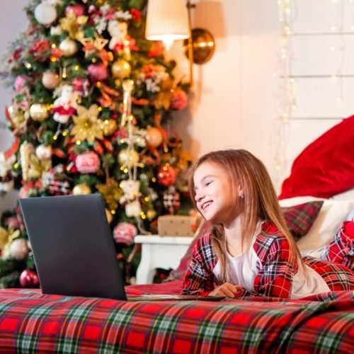 girl in pajamas lies on a bed on red plaid in the background of a Christmas tree and looks at the monitor of the laptop.